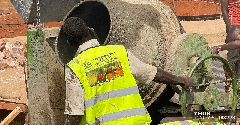 A site worker operating a concrete mixer at a construction site. The worker is wearing a reflector branded by YHDR builders Uganda.